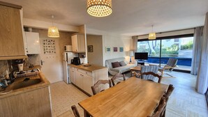 A modern kitchen with wooden cabinets, a dining table, and a sofa. There is a large window with a view of a pool and greenery. - 2 rooms, WIFI, sea view, garden, private parking (ST MANDRIER SUR MER)