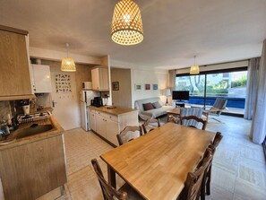 A modern kitchen with wooden cabinets, a dining table, and a sofa. There is a large window with a view of a pool and greenery. - 2 rooms, WIFI, sea view, garden, private parking (ST MANDRIER SUR MER)
