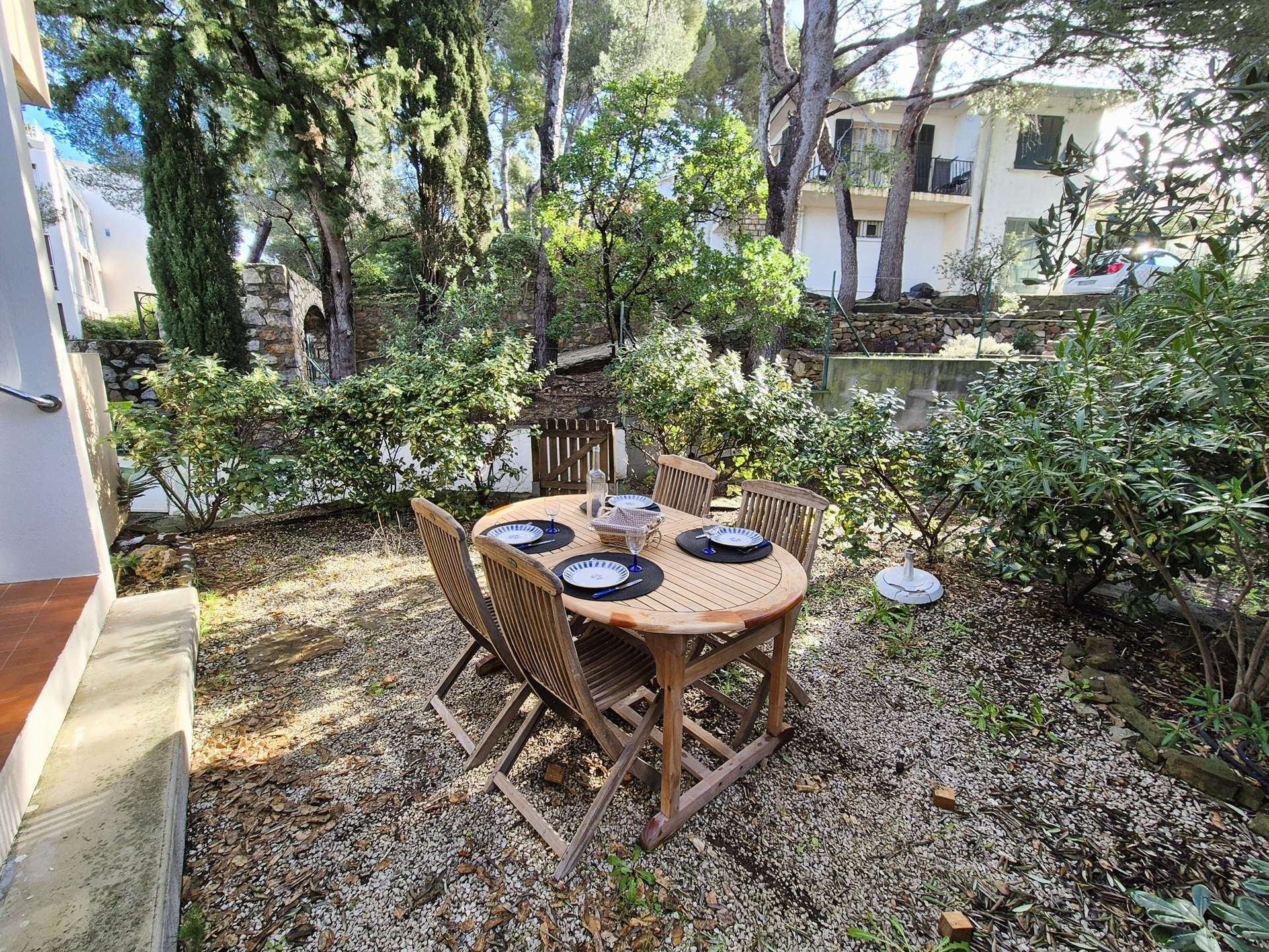 An outdoor dining area with a round wooden table and chairs, surrounded by trees and a stone wall.