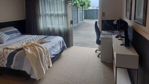 A bedroom with a bed, a desk, and a computer. There is a window with curtains, and a door leading outside. - Tasman Tranquility Cozy family home (Motueka)