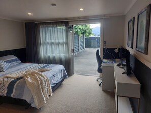 Ein Schlafzimmer mit einem Bett, einem Schreibtisch und einem Computer. Es gibt ein Fenster mit Vorhängen und eine Tür, die nach draußen führt.