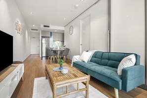 A modern living room with a sofa, coffee table, and TV. There is a dining area with chairs and a table in the background. - 2BR CBD Zen Retreat • Pool, Gym & Free Parking (Melbourne)
