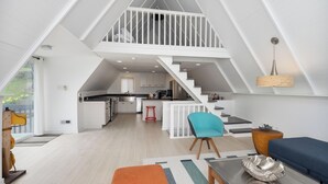 Ampla área de estar estilo loft, com cozinha, escada para um andar superior e vista para o verde lá fora.