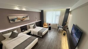Ein Hotelzimmer mit zwei Betten, einem Schreibtisch, einem Sessel, einem Fernseher und einem großen Fenster mit Vorhängen.