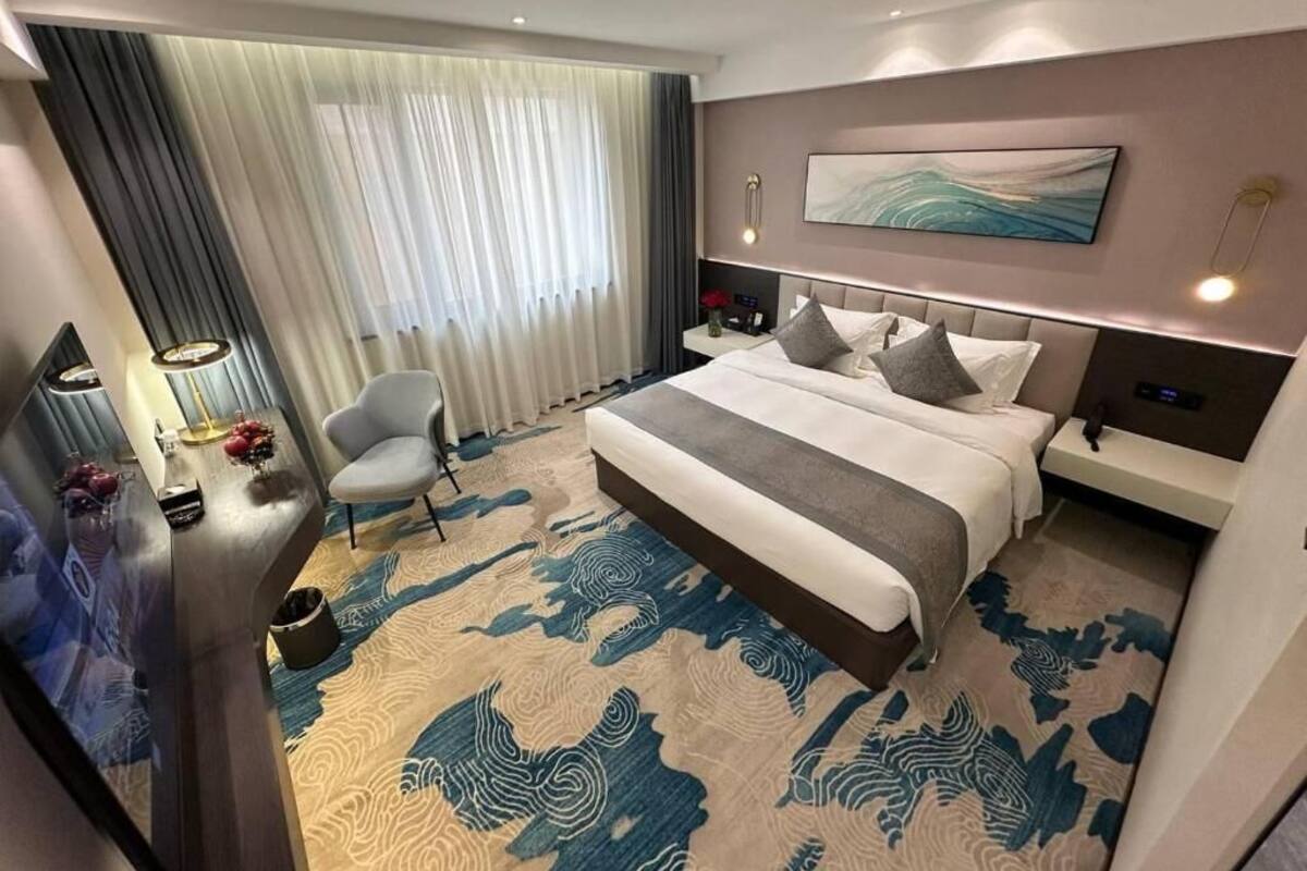 Ein modernes Hotelzimmer mit einem großen Bett, einem Schreibtisch mit Lampe, einem Sessel und Blick auf die Stadt durch das Fenster.