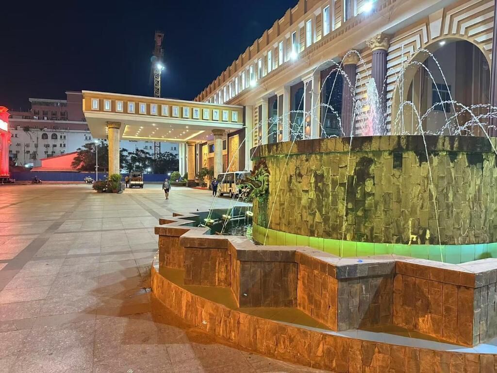 Una plaza pública bien iluminada con una fuente, un corredor cubierto y un edificio de arquitectura clásica.