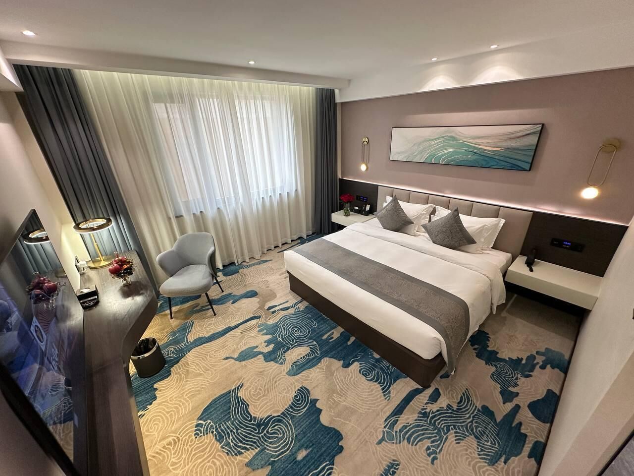 Una habitación de hotel moderna con una cama grande, un escritorio con lámpara, una silla y un ventanal con cortinas.