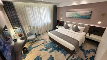 Una habitación de hotel moderna con una cama grande, un escritorio con lámpara, una silla y un ventanal con cortinas.