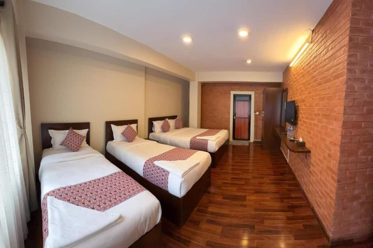 A hotel room with three beds aligned against a brick wall, wooden flooring, and a television on the wall.