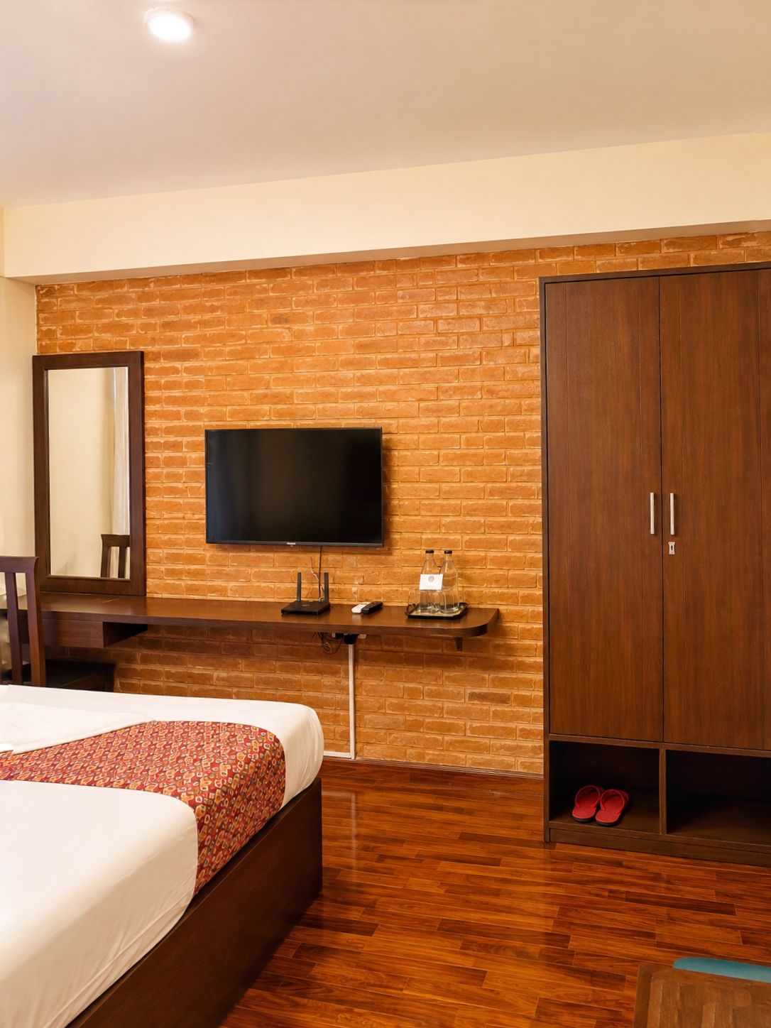 Een hotelkamer met een bakstenen muur, een flatscreen televisie, een houten garderobe, een bed met beddengoed met een patroon, en een paar rode slippers.