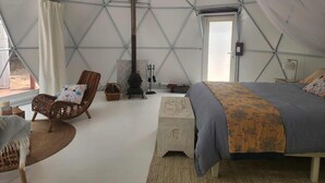 A tented room with a bed, a chair, a small table, a basket, and a fireplace. - Solara Dome Off Grid Luxury and Stillness at Shanlyn Farm (Traveston)