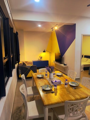 A dining area with a wooden table set for four, surrounded by white chairs. There is a sofa and a TV in the background. - 10 mins from KLCC cozy place to stay (Kuala Lumpur)