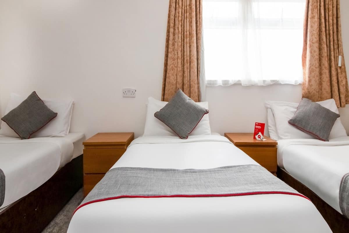 Habitación de hotel con dos camas individuales, una mesita de noche de madera, un objeto rojo sobre la mesita y una ventana con cortinas.