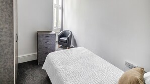 A bedroom with a bed, a chair, a dresser, and a window. - Standard Quad Room (Salford)