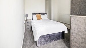 A bedroom with a bed, a nightstand, a lamp, and a wardrobe. - Standard Quad Room (Salford)