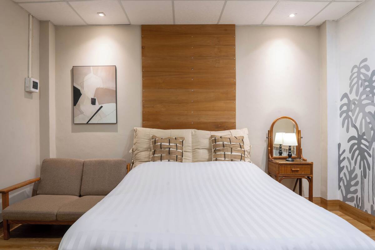 A bedroom with a large bed, a wooden headboard, a bedside table with a lamp, a chair, and a wall-mounted artwork.