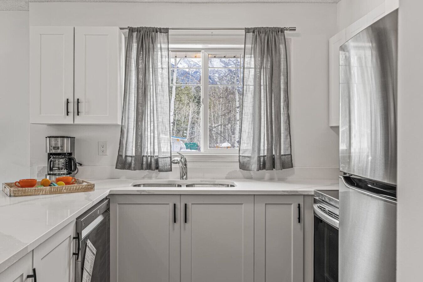 Una cocina con armarios blancos, un refrigerador de acero inoxidable, una cafetera y una ventana con cortinas grises.