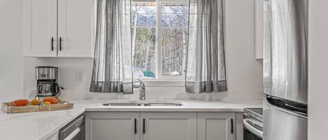 Una cocina con armarios blancos, un refrigerador de acero inoxidable, una cafetera y una ventana con cortinas grises.