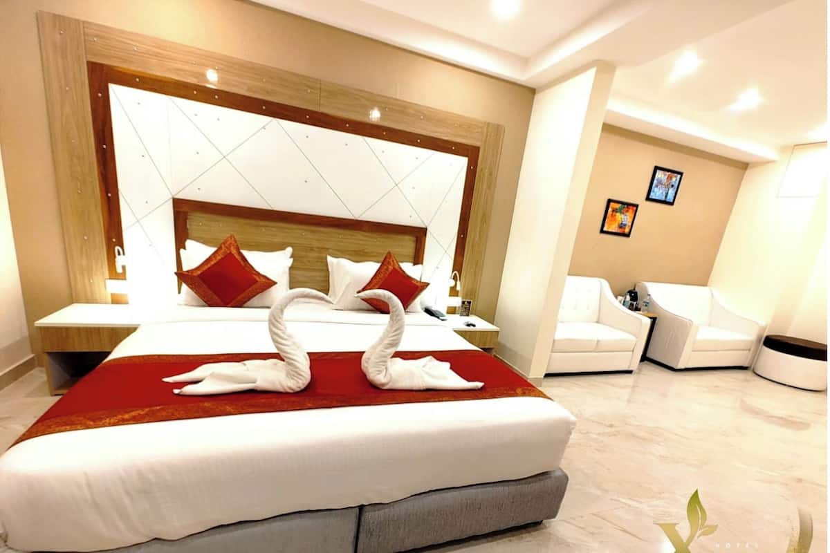 Una habitación de hotel con una cama grande, cabecera de madera, una manta roja y dos toallas con forma de cisne. Hay un sillón blanco y una mesita con una lámpara.