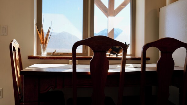 Una scrivania in legno con due sedie, una finestra che si affaccia su montagne e un vaso con piante essiccate sul davanzale.