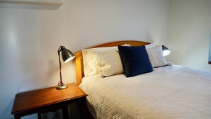 Un letto rifatto con copriletto bianco trapuntato, una testiera in legno e due cuscini, uno bianco e uno blu scuro. Una lampada da scrivania con paralume metallico è posizionata su un comodino in legno accanto al letto.