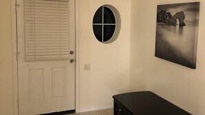 Um quarto com porta branca, janela redonda, um banco preto e luminária no teto.