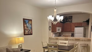 Uma sala de estar com sofá, uma mesa de jantar com cadeiras, um candelabro e uma cozinha com armários de madeira e eletrodomésticos.