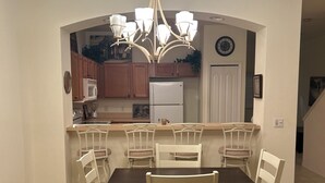 Uma área de refeições com uma mesa de madeira escura e cadeiras brancas, um candelabro e uma cozinha ao fundo.