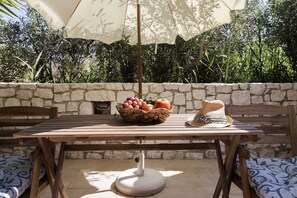 A wooden table with a basket of fruit, a hat, and an umbrella in a shaded outdoor area with stone walls and greenery. - Azalea Villas (Lefkada)
