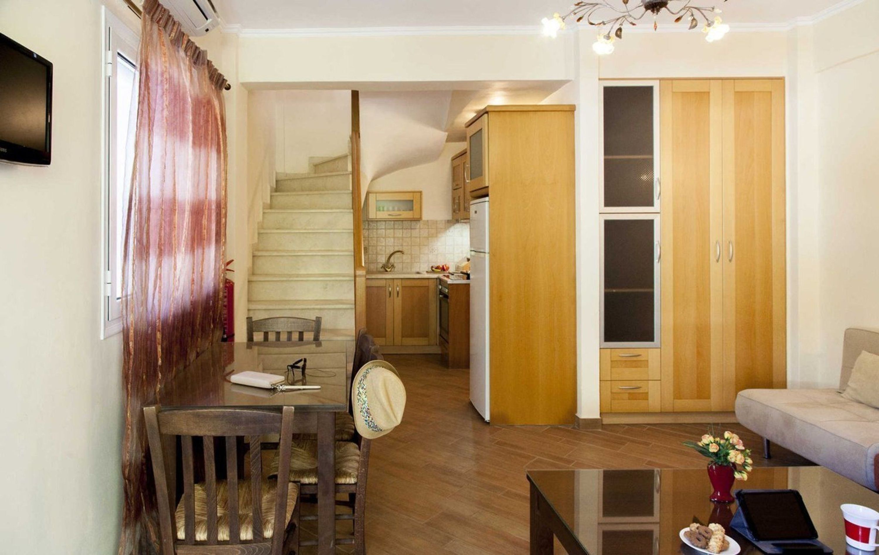 A living room with a sofa, a coffee table, and a dining area with a chair and table. There is a kitchen area with cabinets and a refrigerator. A staircase leads to another level.