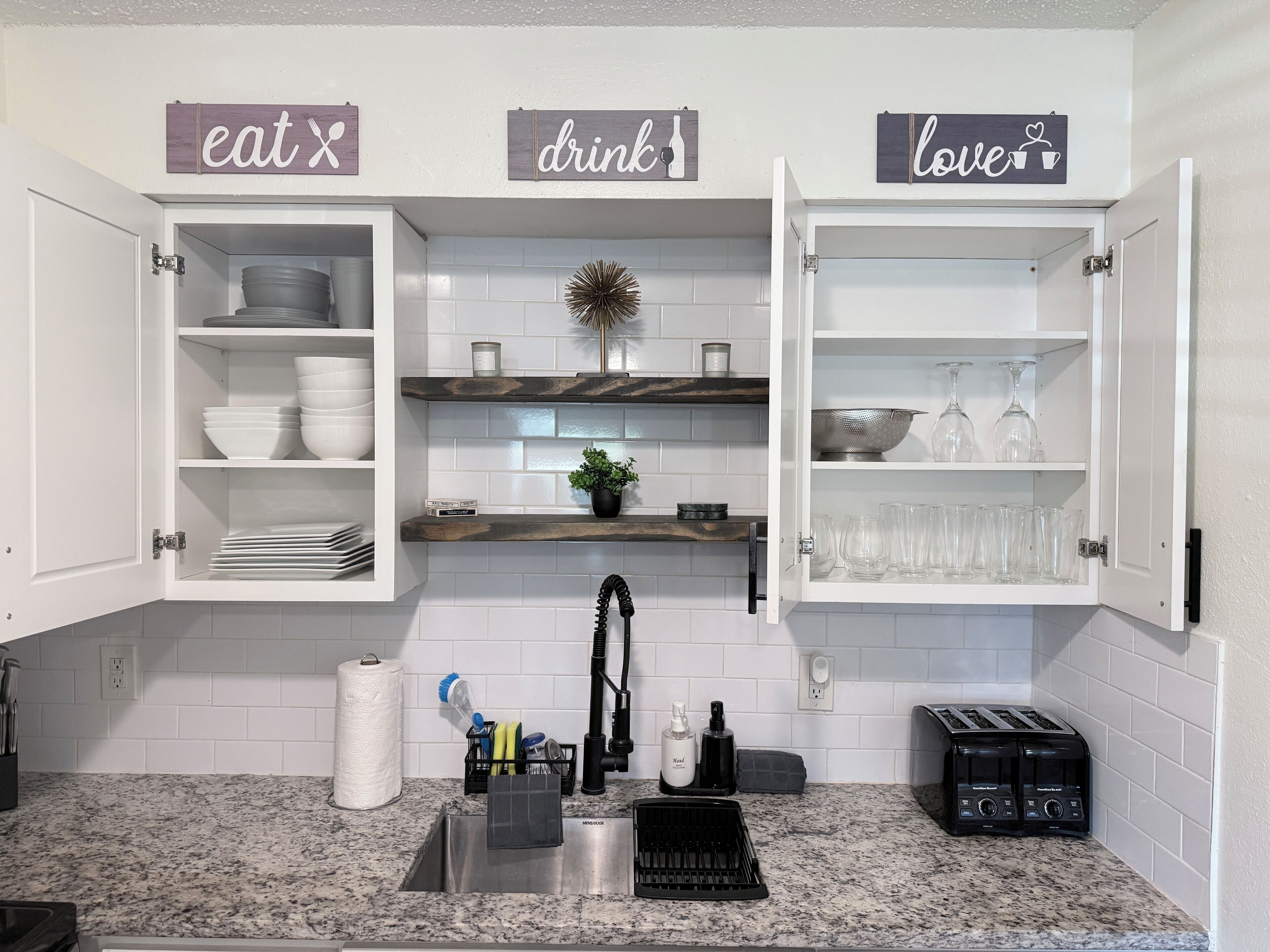 A kitchen with open cabinets displaying dishes, a sink with a faucet, and a countertop with a coffee maker and toaster.