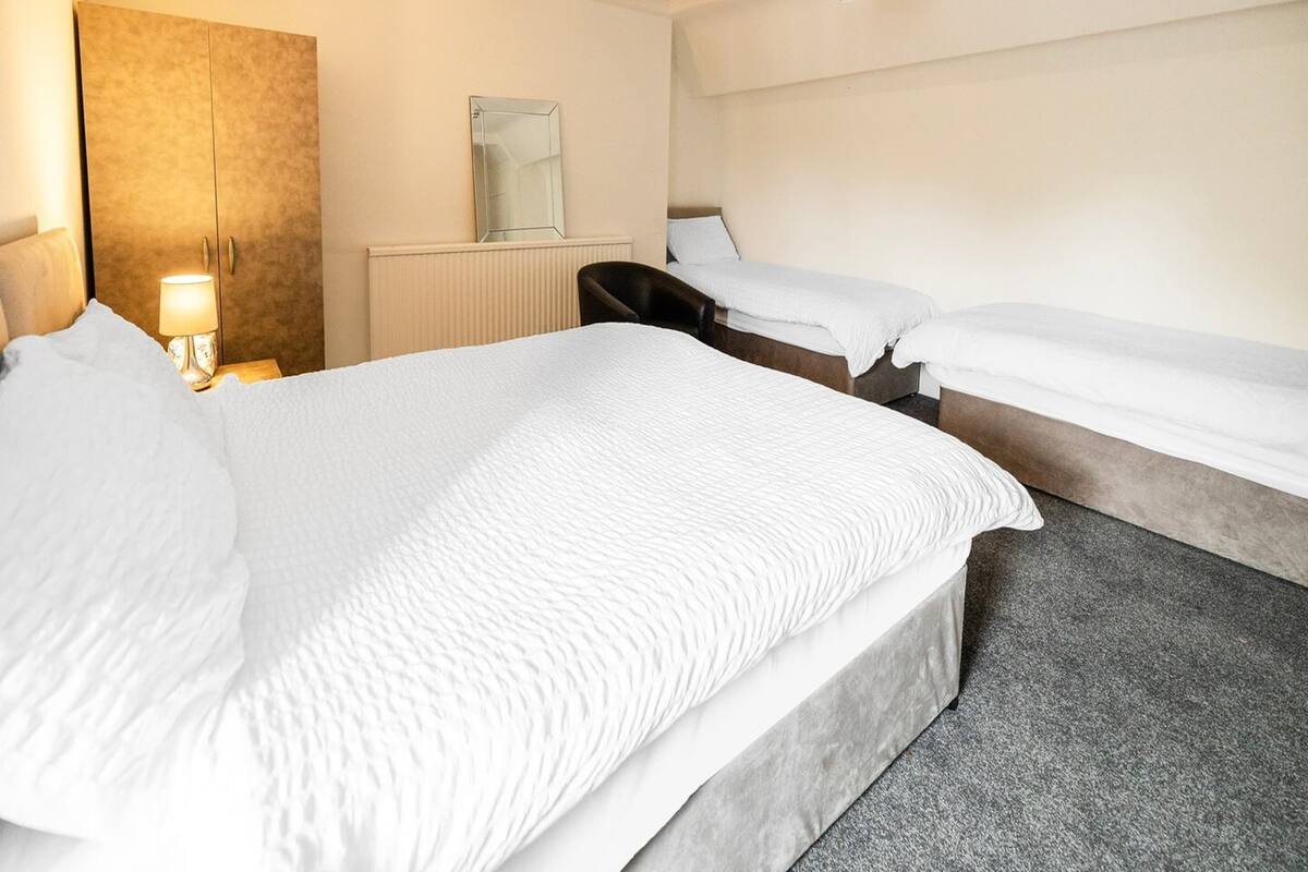 A hotel room with a large bed, two smaller beds, a desk with a lamp, a mirror, and a radiator.