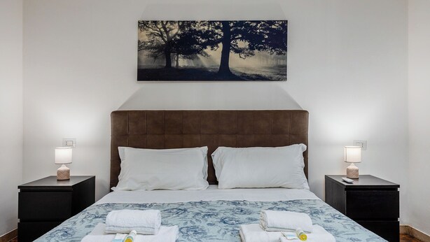 Una cama bien hecha con una colcha estampada con motivos florales, dos almohadas blancas y dos mesitas de noche con lámparas.