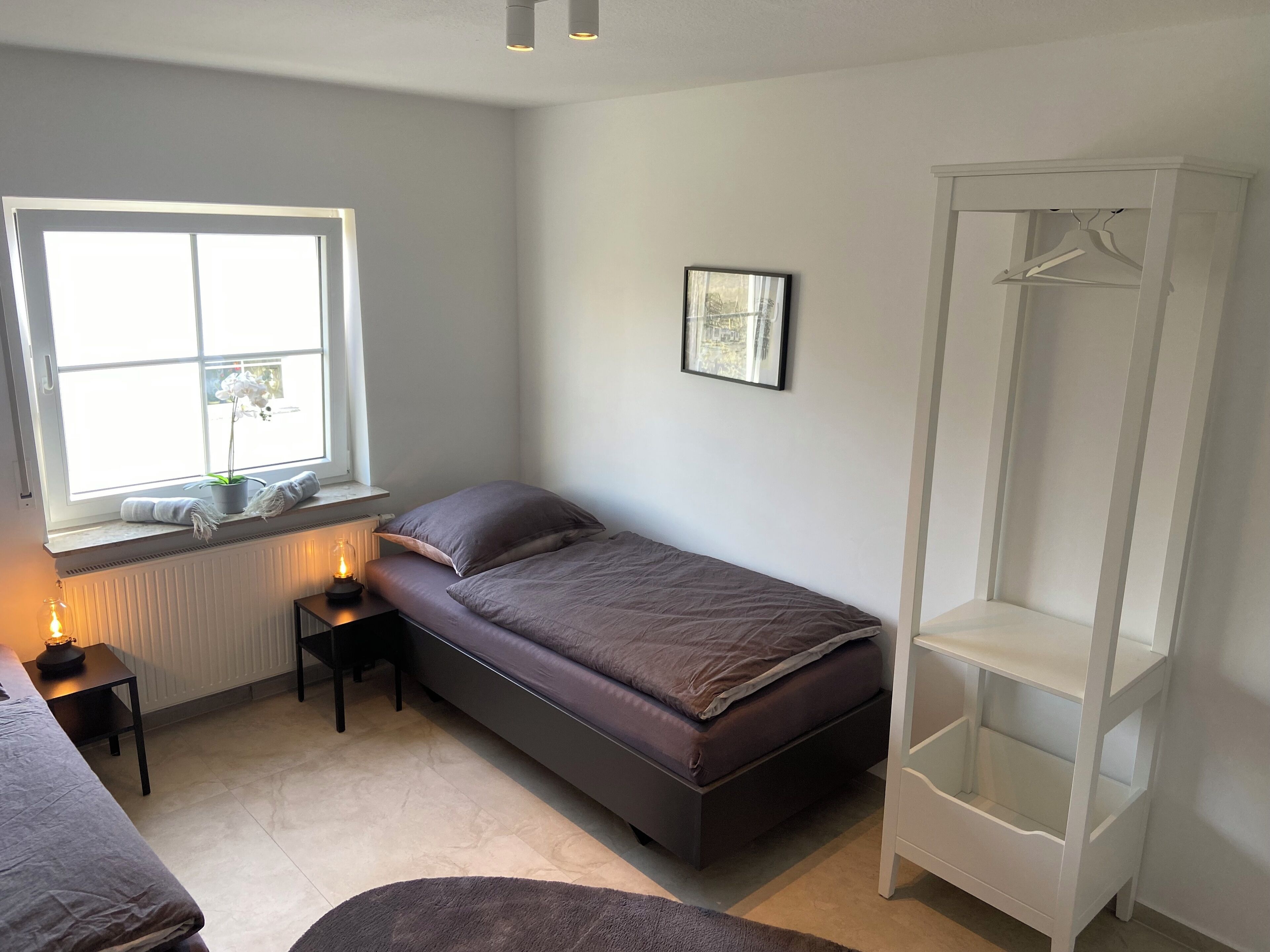 Ein kleines, ordentlich eingerichtetes Schlafzimmer mit einem Einzelbett, einem Nachttisch mit Lampe, einer Heizung, einem Fenster mit Blick und einem weißen Kleiderschrank.