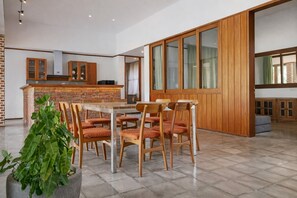 A spacious dining area with a large wooden table and chairs, a brick accent wall, and a view through a large window with wooden paneled walls. - Matthew Villa Umalas (Kerobokan)