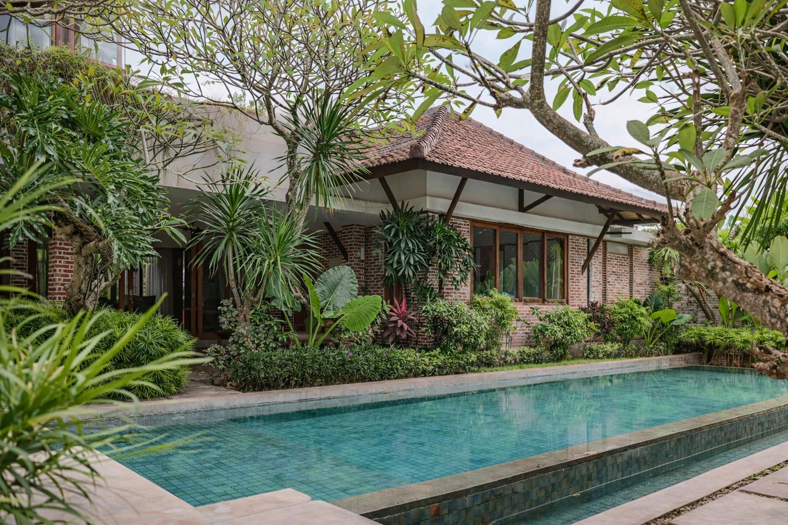 A poolside area with a brick building, surrounded by lush greenery and a tiled swimming pool.