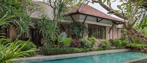 A poolside area with a brick building, surrounded by lush greenery and a tiled swimming pool.