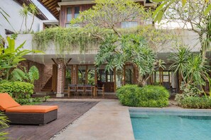 A modern outdoor pool area with a wooden deck, a seating area with a table and chairs, and lush greenery including various plants and trees. - Matthew Villa Umalas (Kerobokan)
