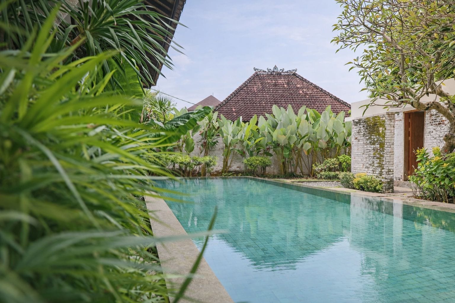 A swimming pool with a tiled deck, surrounded by lush greenery and a traditional building with a tiled roof.