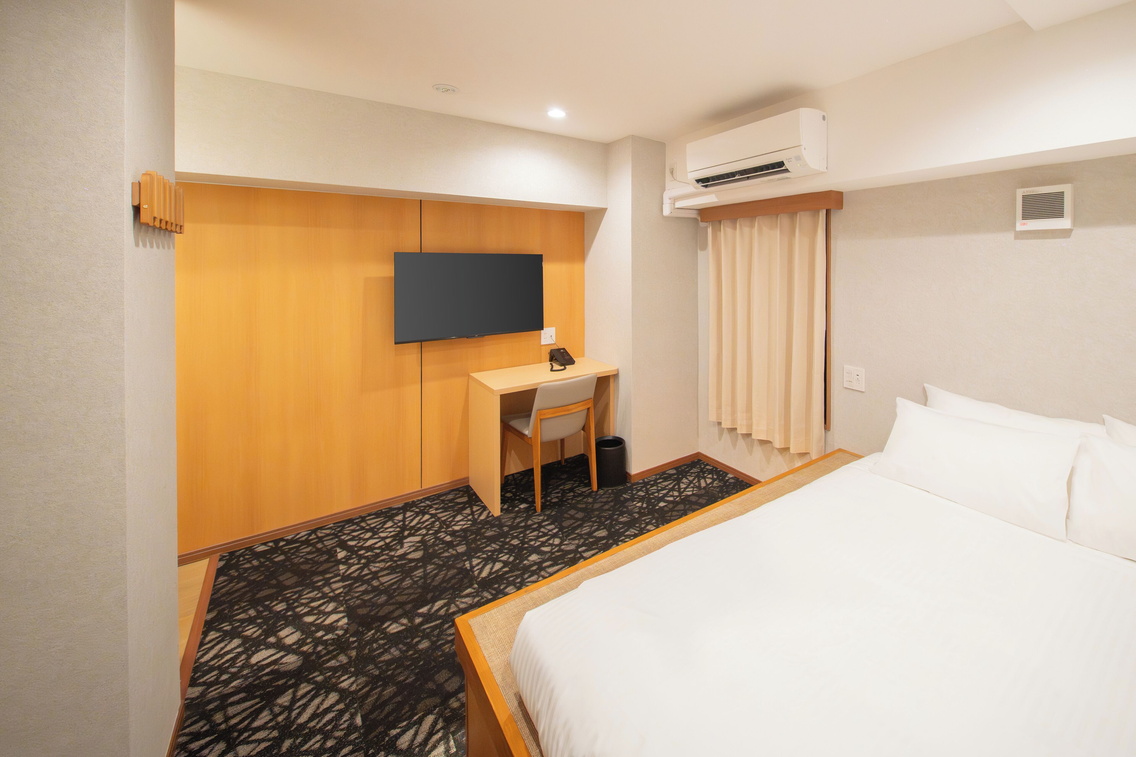 Een hotelkamer met een bed, een bureau met een stoel, een televisie, een airconditioning en een kamerbreed tapijt met dessin.