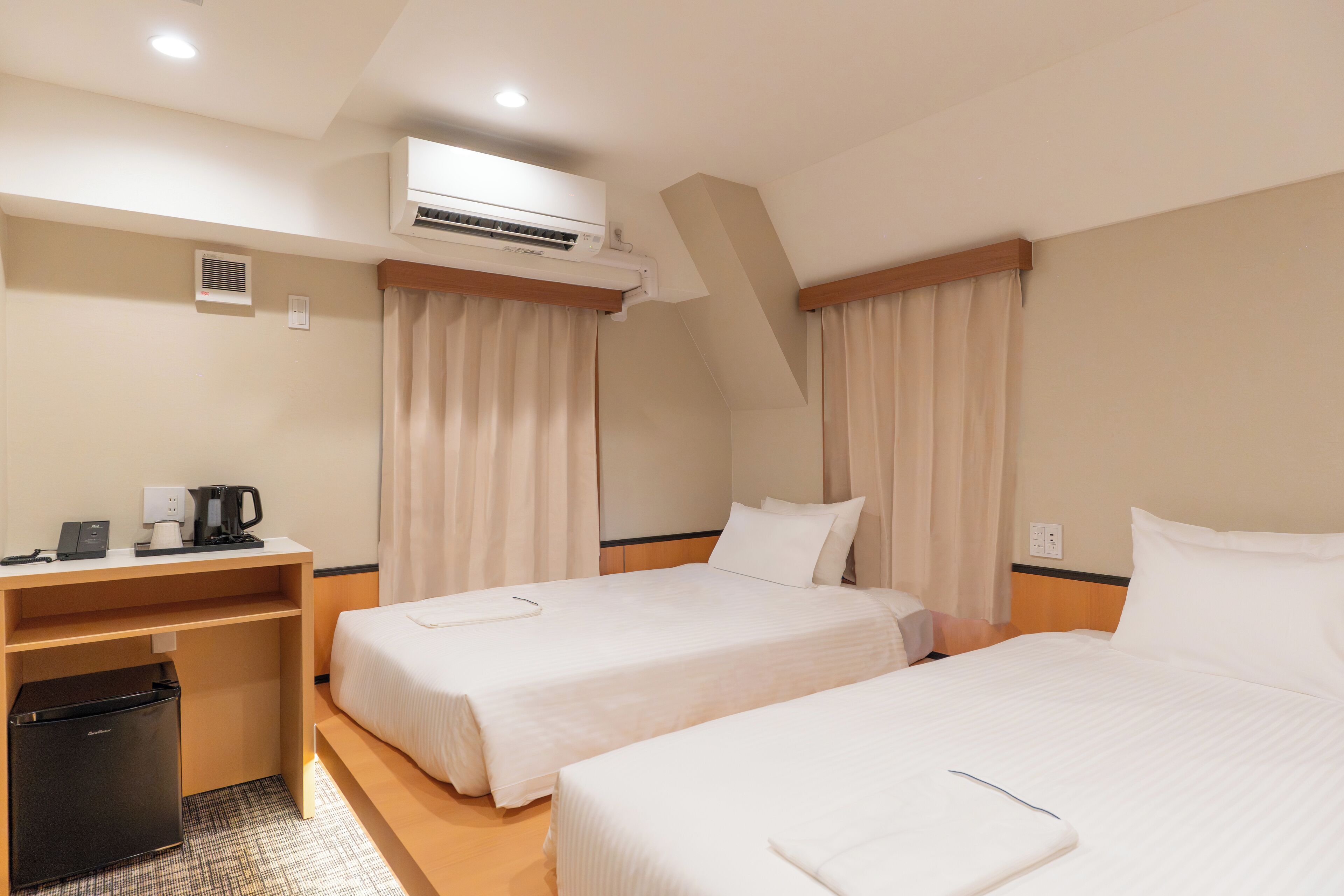 A hotel room with two beds, a desk, a refrigerator, an air conditioner, and curtains.