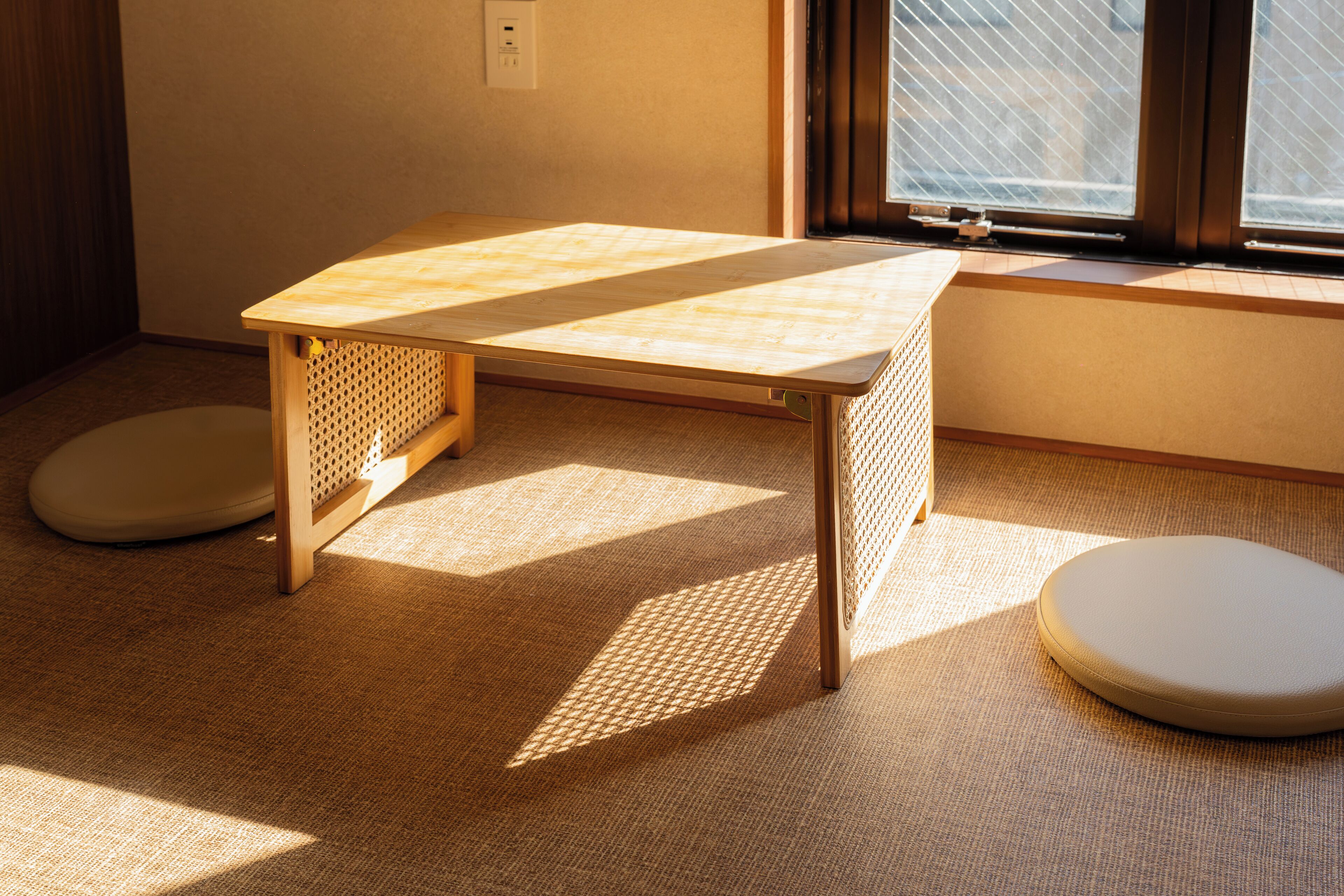 Une pièce avec une petite table en bois, deux tabourets ronds et une fenêtre avec des stores.