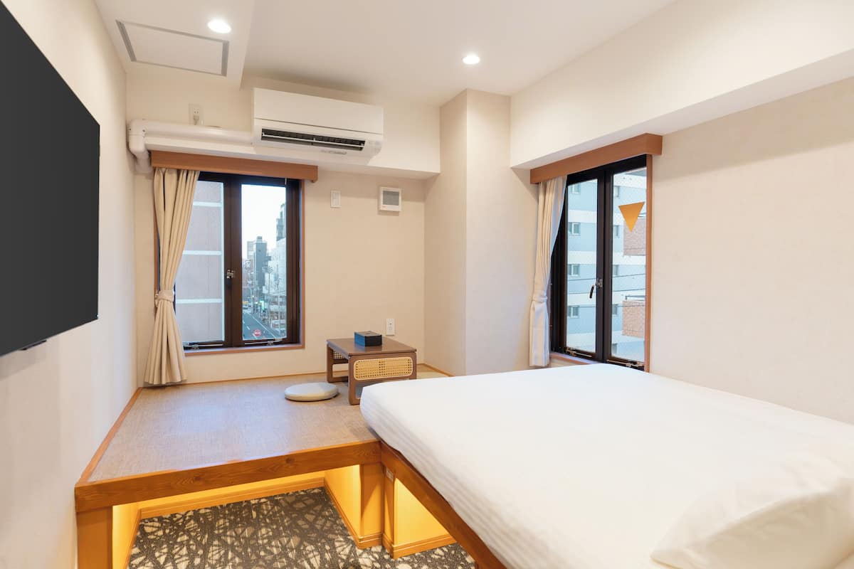 Una camera d'albergo con un letto, un tavolino, una sedia, un condizionatore d'aria e una finestra con tende.