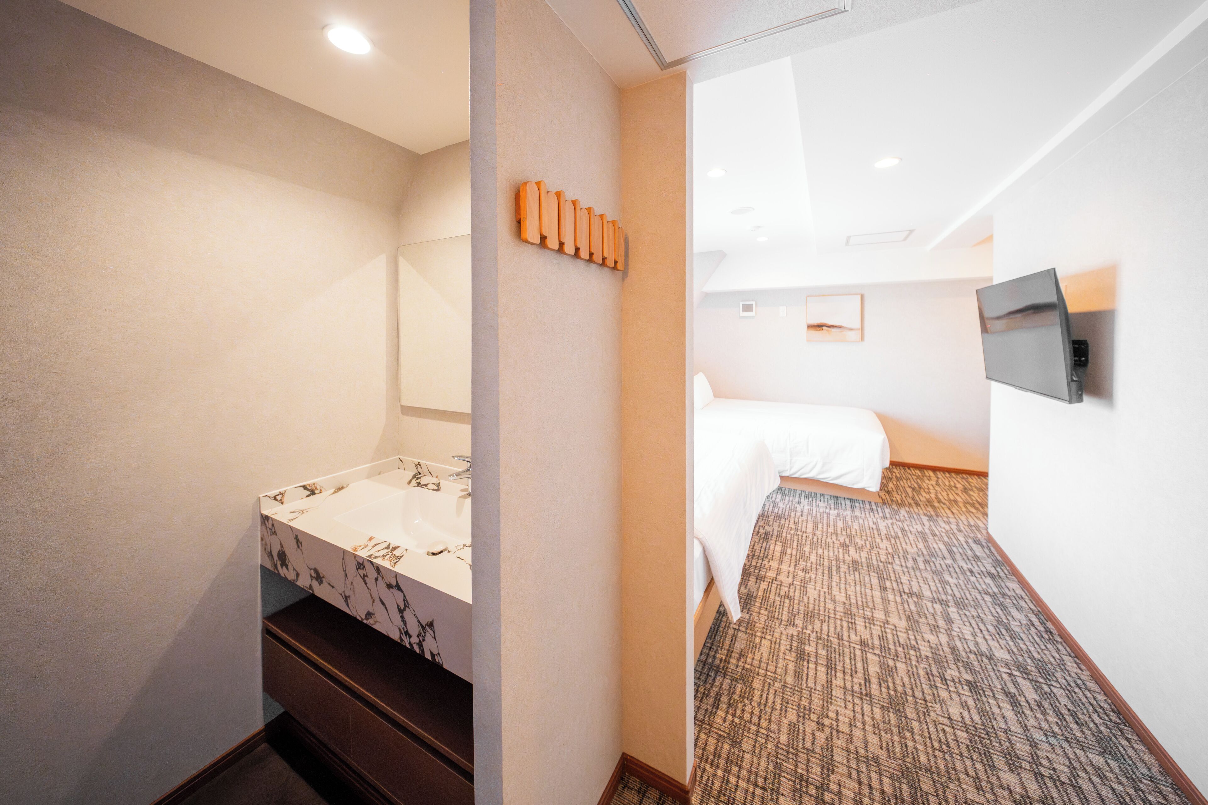 A hotel room with a marble bathroom sink, a bed with white bedding, a television mounted on the wall, and a carpeted floor.