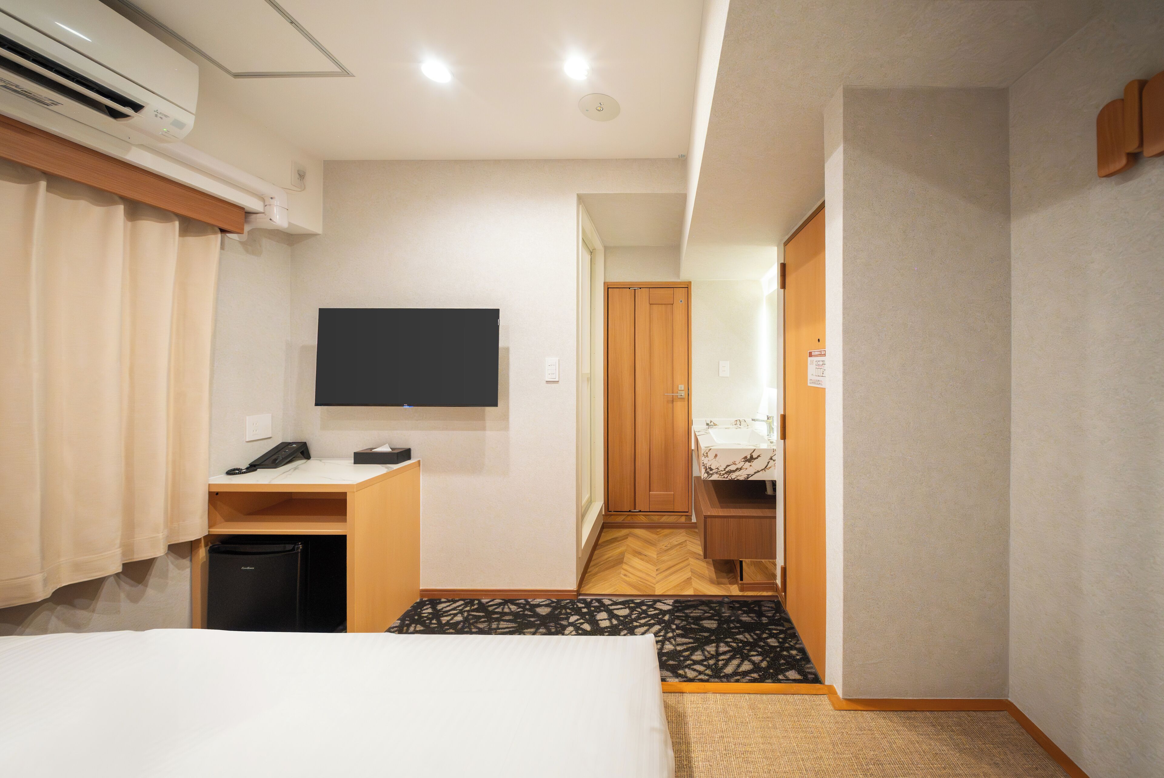 A hotel room with a bed, a desk with a telephone, a flat-screen TV mounted on the wall, a small black trash bin, and a wooden wardrobe.