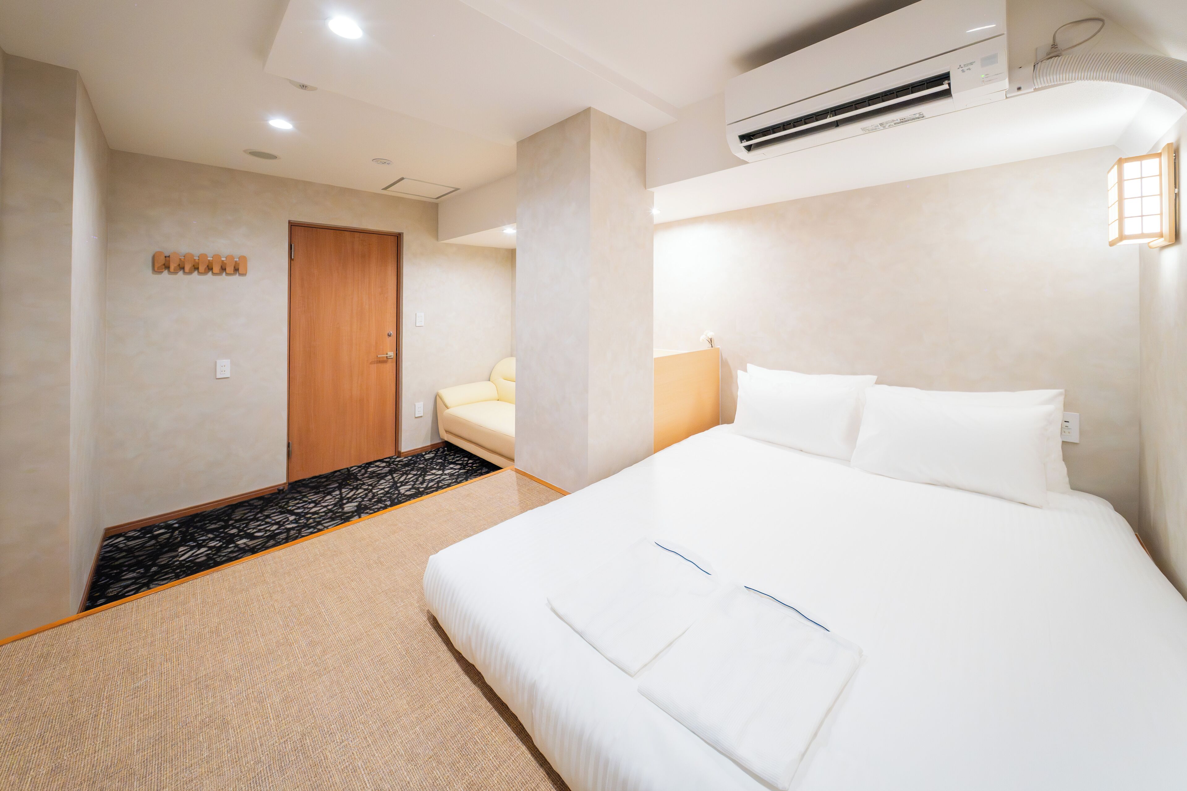 A hotel room with a large bed, a wooden door, a small seating area, and a wall-mounted air conditioner.