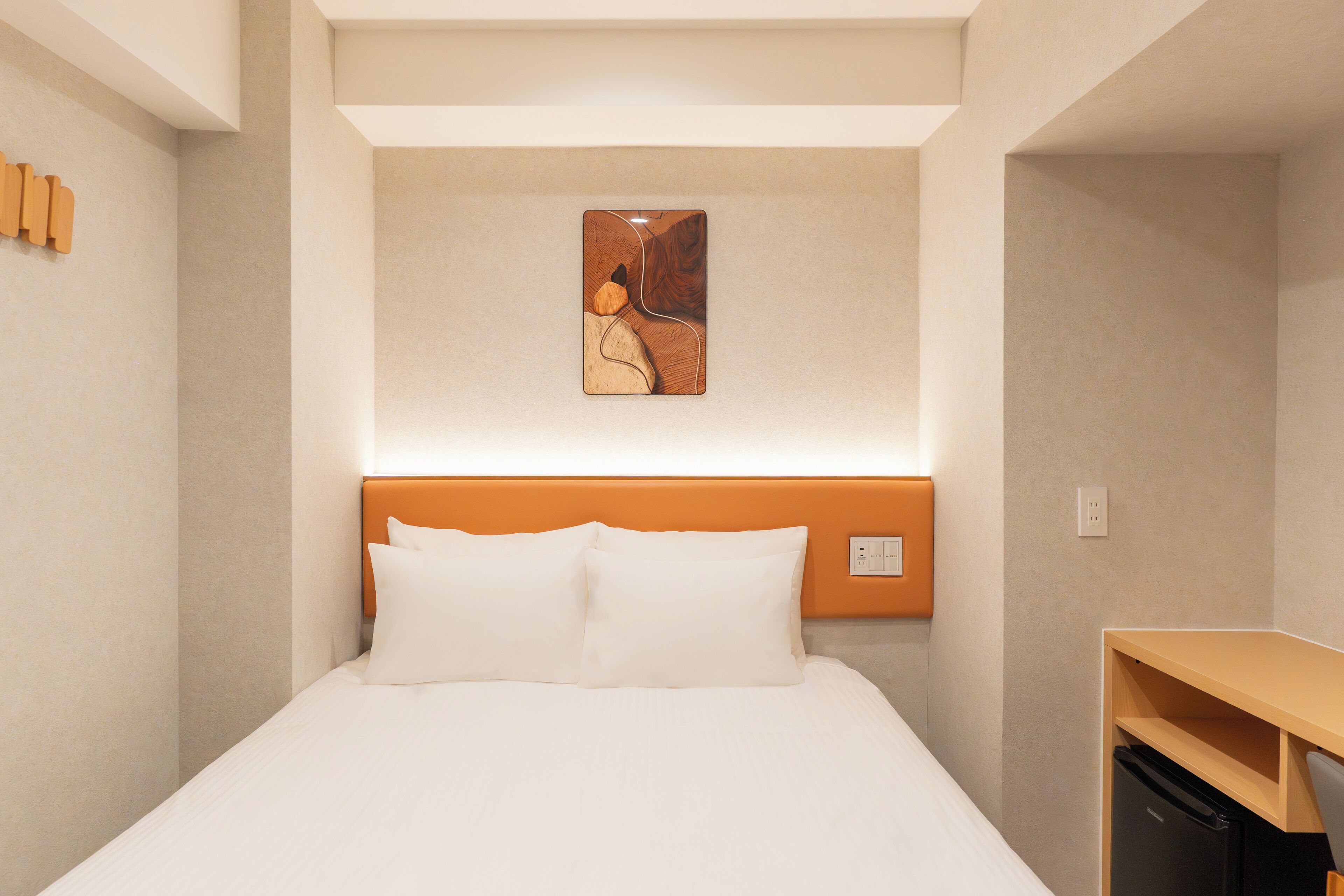 A hotel room with a bed, a headboard, a wall-mounted artwork, and a desk with a chair.