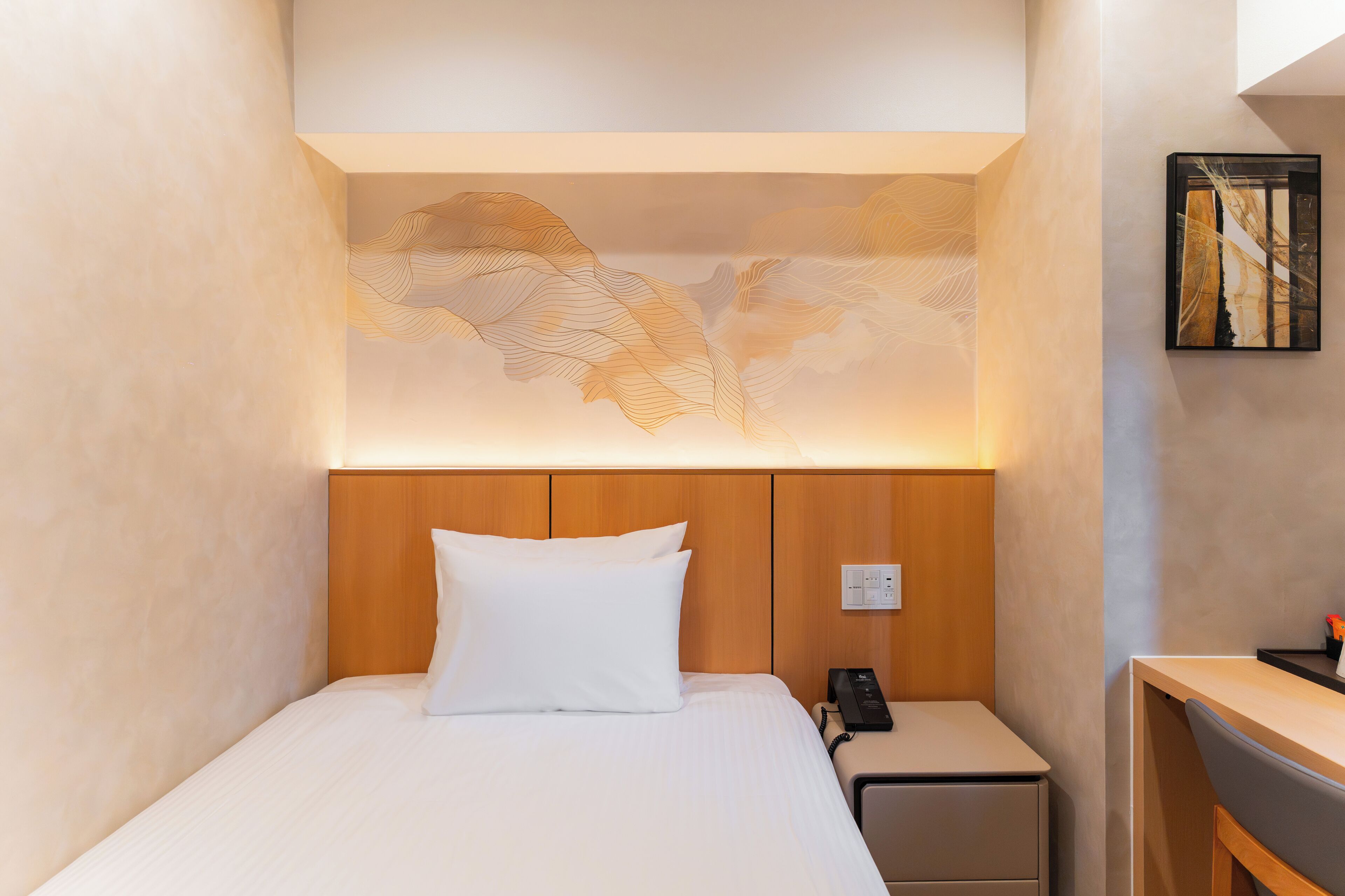 A modern hotel room with a large bed, a wooden headboard, a nightstand with a phone, and a desk with a chair.