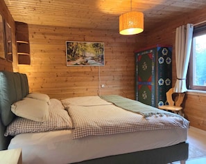 Puinen mökkimainen makuuhuone, jossa on sänky, riippuvalaisin, taulu, kaappi ja ikkuna.