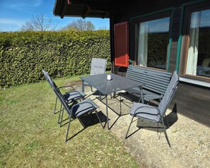 Ein Sitzbereich im Freien mit Tisch und vier Stühlen, ein Hecken im Hintergrund und ein Gebäude mit großen Fenstern.